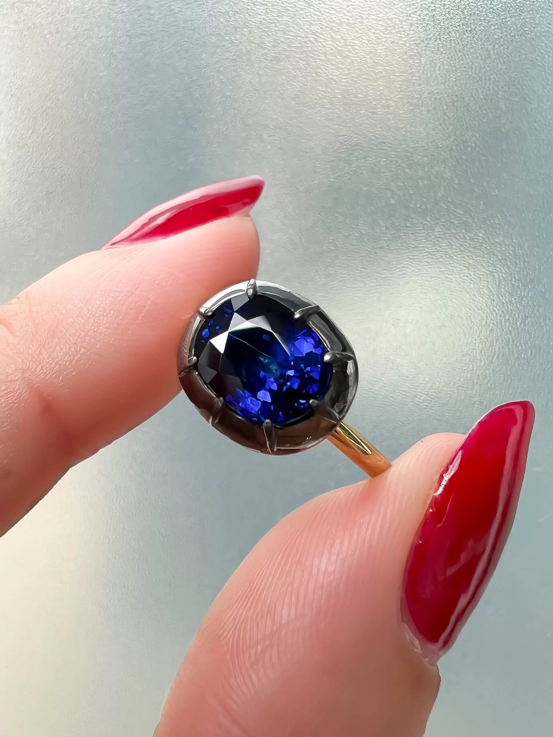 East To West Oval Sapphire Close-up of an East-to-West deep-blue sapphire ring with a black-rhodium Georgian-style setting and yellow-gold band, shown on the finger in natural light.